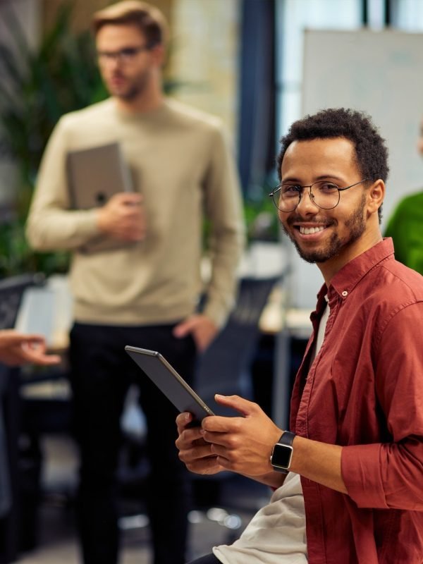 young-cheerful-mixed-race-man-holding-digital-tablet-and-smiling-at-camera-while-working-with